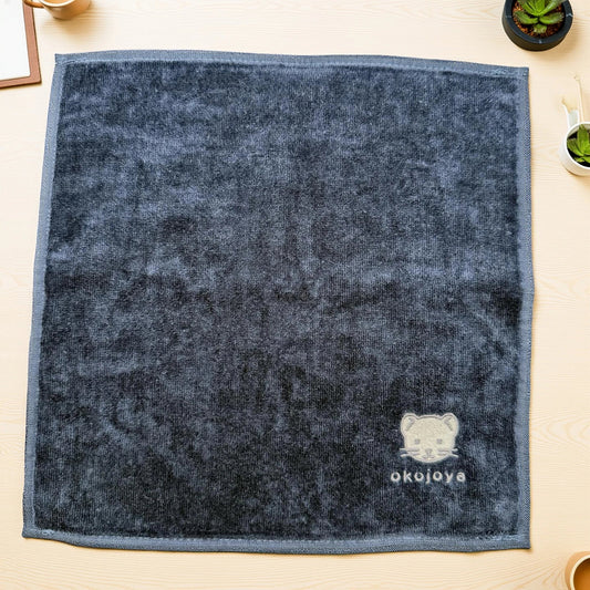 木の机に置いたokojoyaロゴ刺繍ハンカチタオル。観葉植物と並べたギフト風の生活シーン（おこじょ／オコジョ）